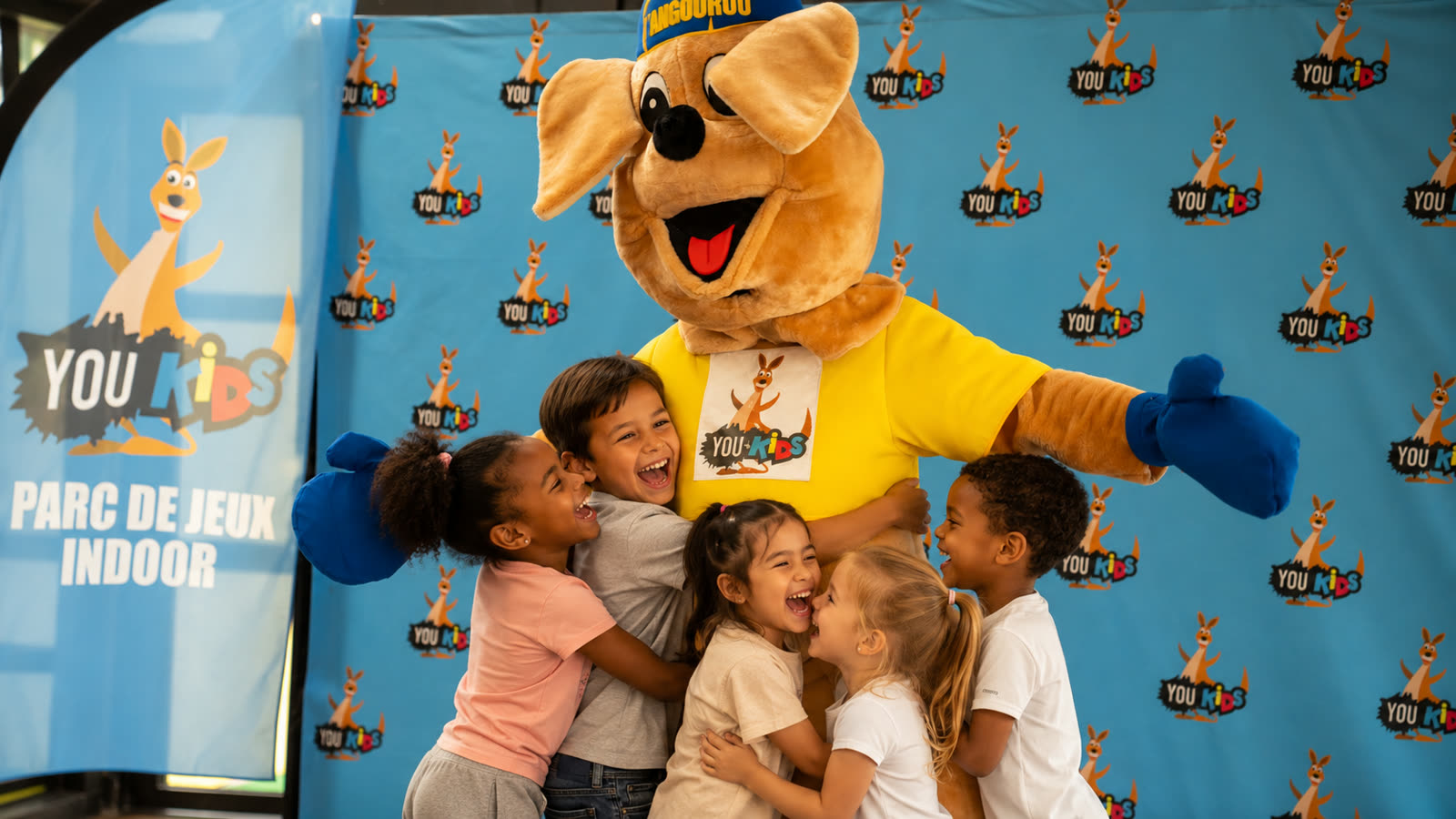 Groupe d'enfants faisant un câlin à la mascotte You Kids lors d'un anniversaire à Thiverval Liberty